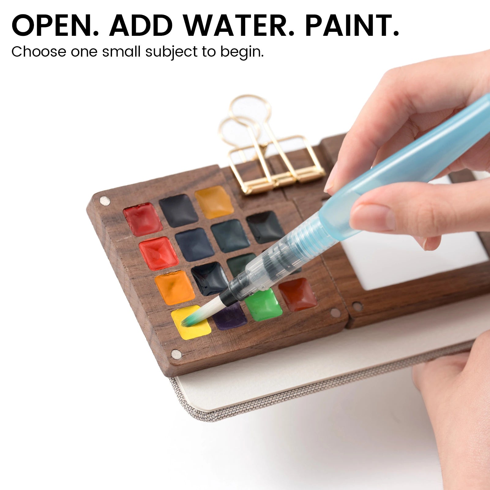 Water brush touching yellow paint in the wooden EXPLORER palette clipped to a journal