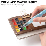Water brush touching yellow paint in the wooden EXPLORER palette clipped to a journal
